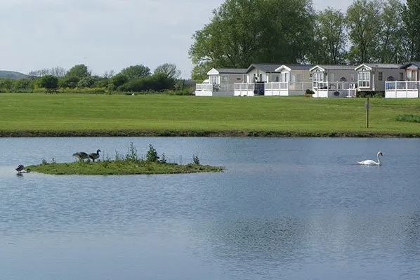 A lake in the foreground with fields and holiday homes in the background