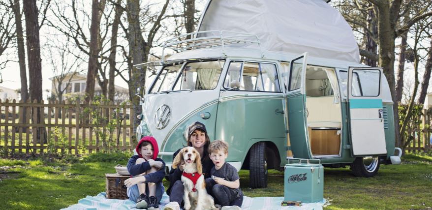 A lady and two boys sitting with their dog on a blanket on the grass in front of a VW camper