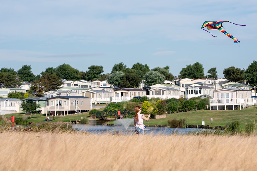 A child flying a kite in the foreground of a lake and holiday homes in the distance on a bright sunnt day