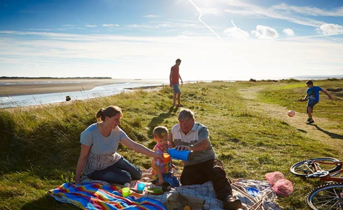 A family sat down on a picnic mat with other people playing football nearby next to an estuary on a bright summers day