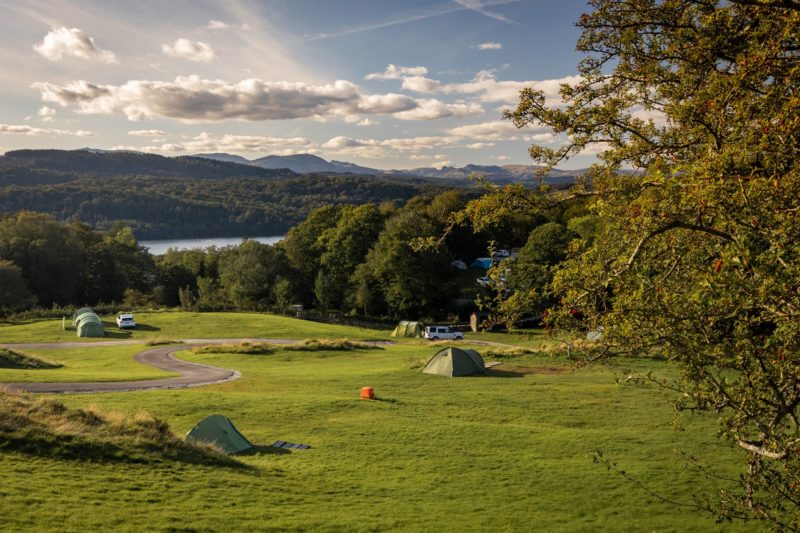 A view of a lake in the far distance and a rolling hill leading down to the shore in amongst trees