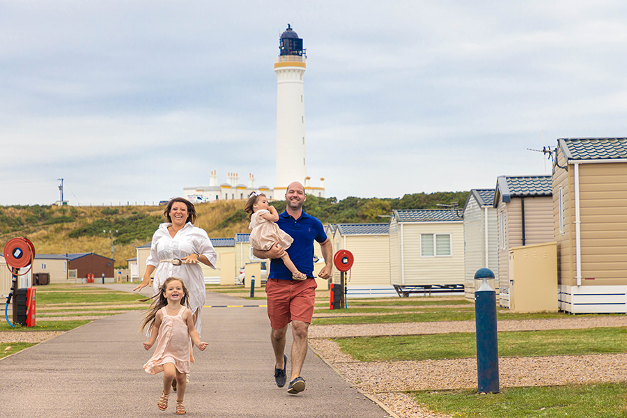A family running towards the camera surrounded by holiday homes with a lighthouse in the distance