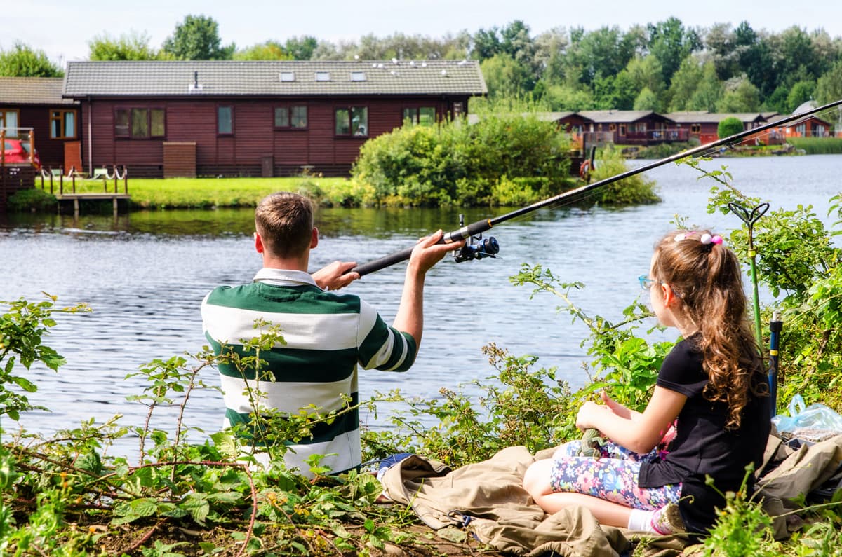 Two people sat next to a lake fishing surrounded by holiday homes