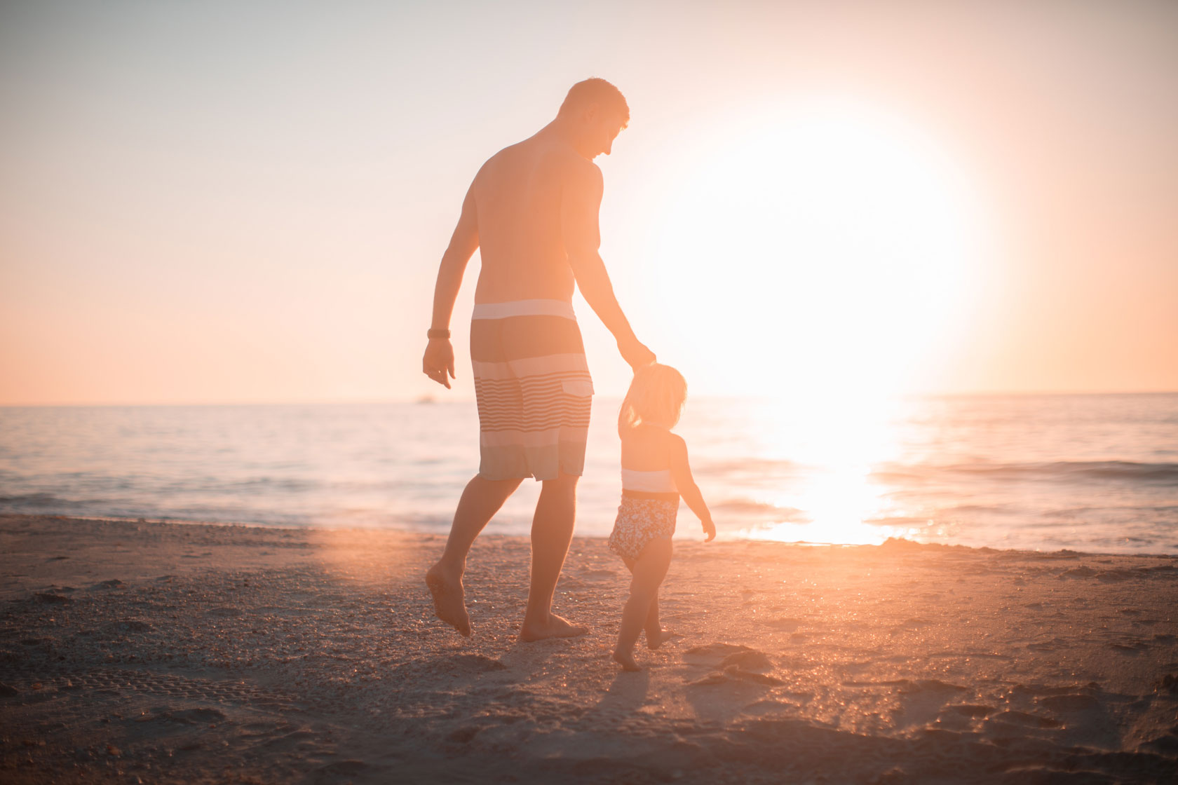 A Adult Male Walking a Small Child Towards the Sea as the Sun is Setting