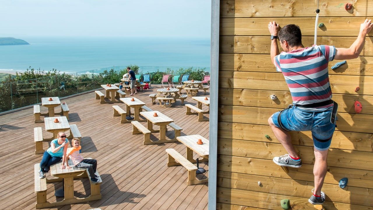 A Parent Climbing a Climbing Wall with Others Watching with Glorious Sea Views in the Background