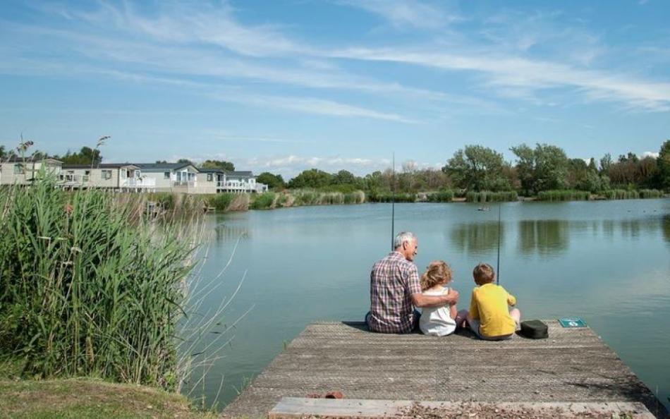 A grand parent sitting with his two grandchildren on a jetty reaching out over a lake fishing