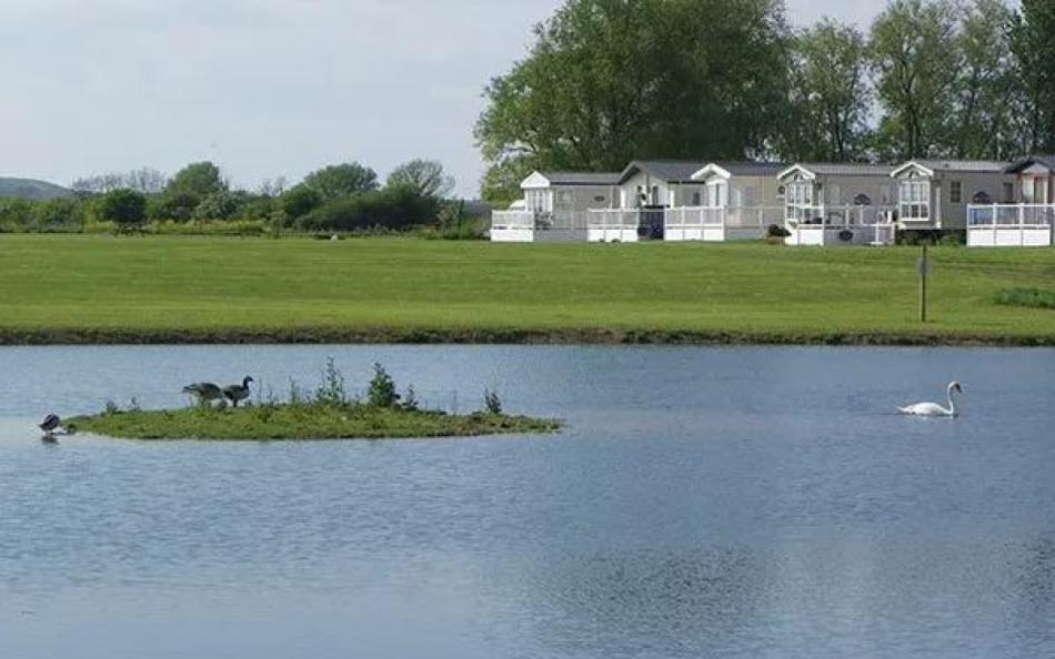 A lake in the foreground with fields and holiday homes in the background