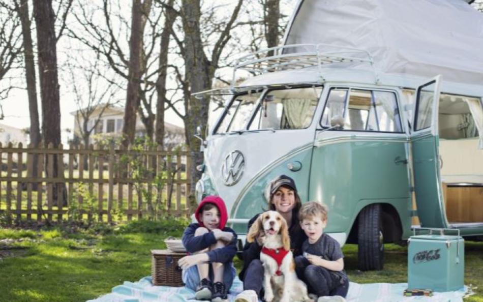 A lady and two boys sitting with their dog on a blanket on the grass in front of a VW camper