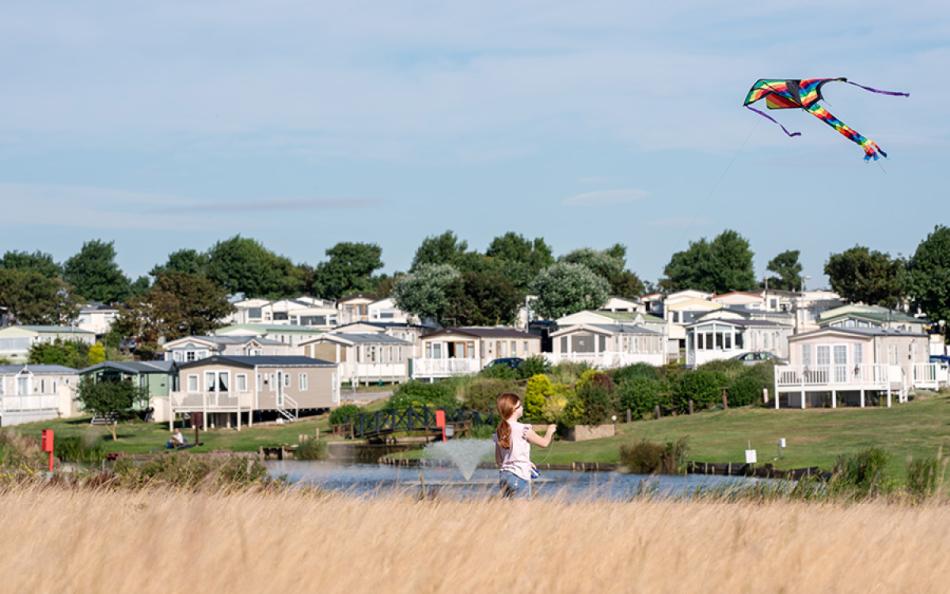 A child flying a kite in the foreground of a lake and holiday homes in the distance on a bright sunnt day