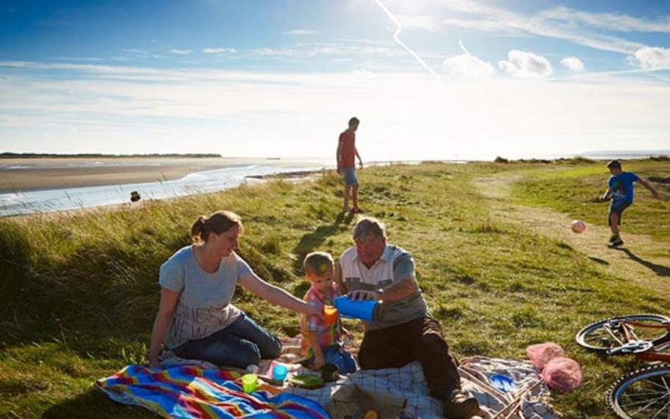 A family sat down on a picnic mat with other people playing football nearby next to an estuary on a bright summers day