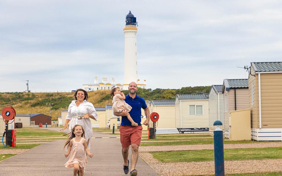 A family running towards the camera surrounded by holiday homes with a lighthouse in the distance