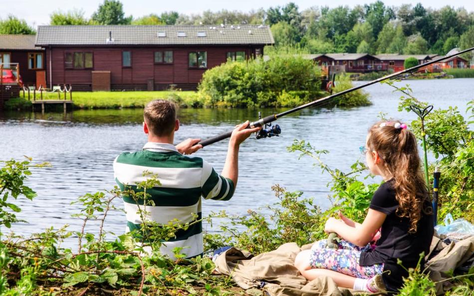 Two people sat next to a lake fishing surrounded by holiday homes
