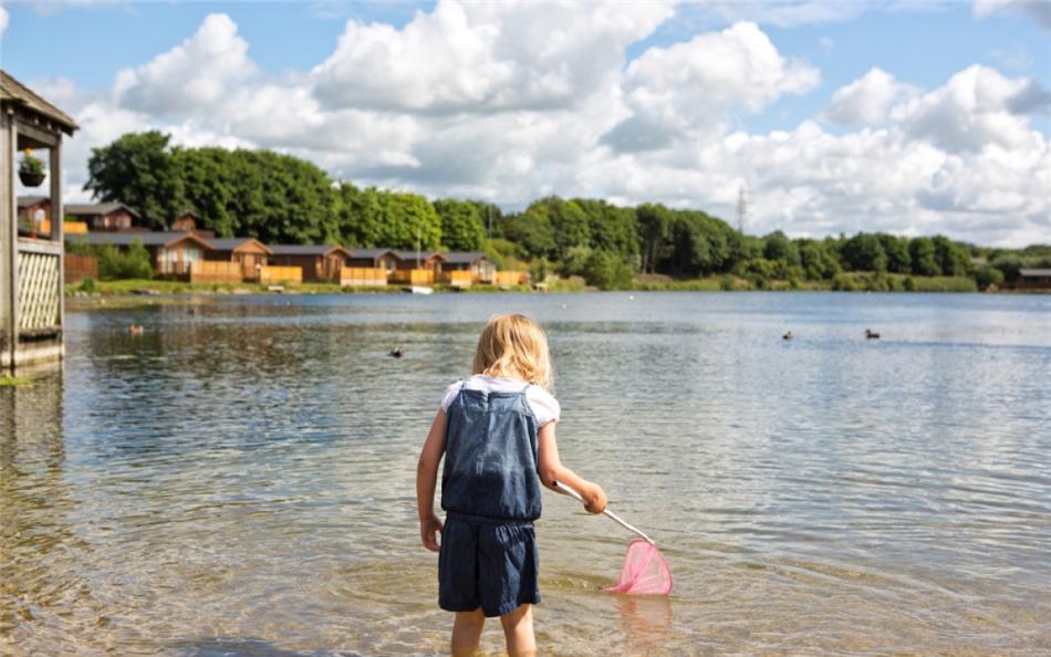 A child fishing with a pink net in the shallows of a lake surrounded by holiday homes and forests