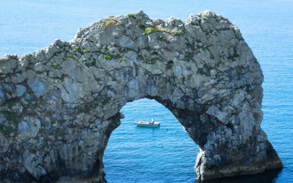  An image of bright blue sea with a small cave looking through to open water with a sailing boat in the background