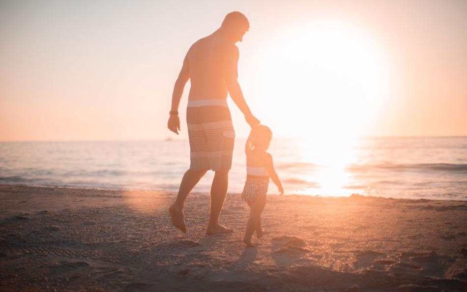 A Adult Male Walking a Small Child Towards the Sea as the Sun is Setting
