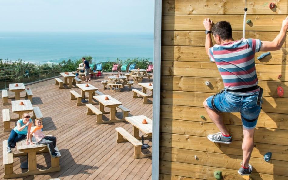 A Parent Climbing a Climbing Wall with Others Watching with Glorious Sea Views in the Background