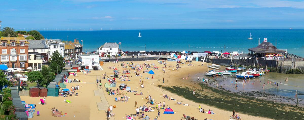 Image of a beach and small harbour with brightly coloured boats and people and sun shades on the beach