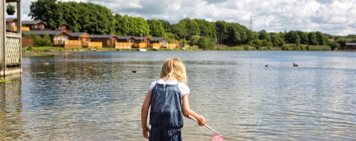 A child fishing with a pink net in the shallows of a lake surrounded by holiday homes and forests